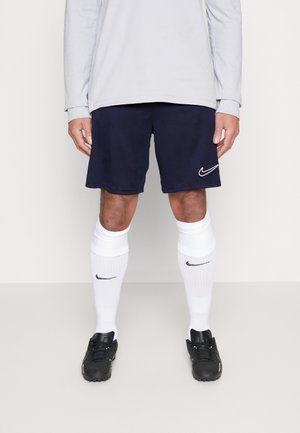 Personne portant un short Nike bleu marine, des chaussettes Nike blanches montantes jusqu'aux genoux, des crampons noirs et un t-shirt à manches longues gris clair, debout sur un sol uni.