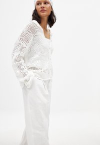 Weiße gehäkelte Strickjacke mit strukturiertem Muster, Knöpfen auf der Vorderseite, kombiniert mit weißen Leinenhosen. Das Outfit wird mit einem passenden gehäkelten Haarband abgerundet.