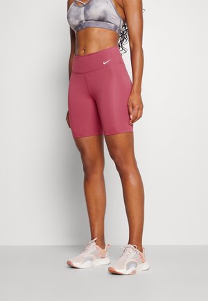 Femme portant un short de vélo Nike mauve, un brassard de sport gris et des baskets de couleur claire, debout contre un fond uni clair.