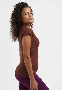 Femme aux cheveux bouclés et foncés, portant un t-shirt marron à manches courtes et des leggings violets, debout de profil sur un fond blanc uni.