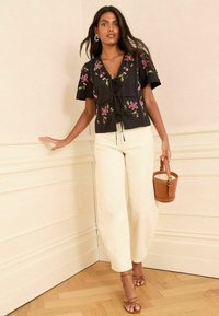 Melnā īsām piedurknēm blūze ar ziedu izšuvumu, sasieta viduklī; kopā ar krēmkrāsas platajām biksēm un brūnu spaiša formādas somu.