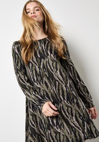 Donna con lunghi capelli biondi che indossa un vestito a maniche lunghe con strisce astratte di tigre nere, oliva e taupe, su uno sfondo chiaro e uniforme.