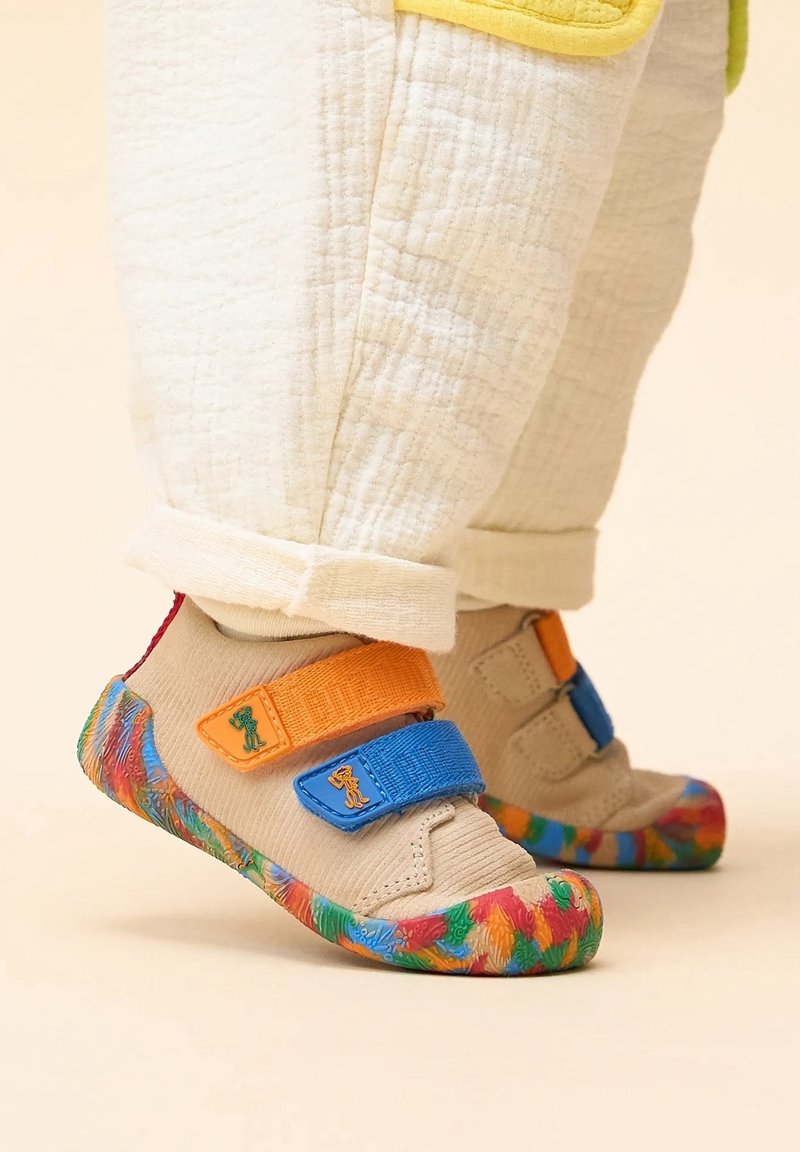 Chaussures marron clair avec semelles à motifs colorés et sangles en velcro bleu et orange. Portées avec un pantalon crème texturé, mettant en valeur un design ludique.