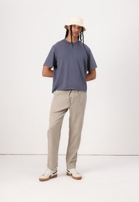 T-shirt gris, pantalon beige à cordon de serrage et chapeau seau beige. Baskets blanches avec semelles marron et accents noirs. Tenue simple et décontractée.