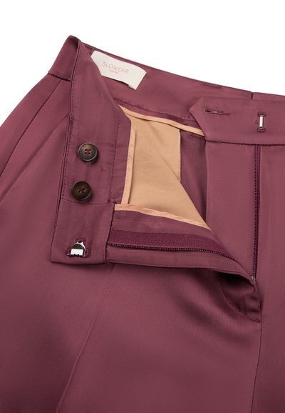Pantalones morados con cremallera abierta, dos botones oscuros, gancho de metal y forro interior beige, mostrando la cintura y la etiqueta de la marca.