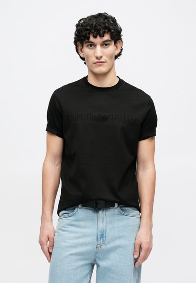 Μαύρο βαμβακερό μπλουζάκι με γυρισμένα μανίκια, με ανάγλυφο λογότυπο "EMPORIO ARMANI" στο στήθος. Συνδυάζεται με ανοιχτόχρωμο τζιν παντελόνι.