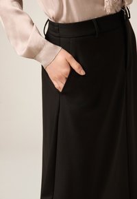 Svarta vida byxor med slät textur, med sidoficka och hög midja, kombinerade med en ljusrosa sidenblus.