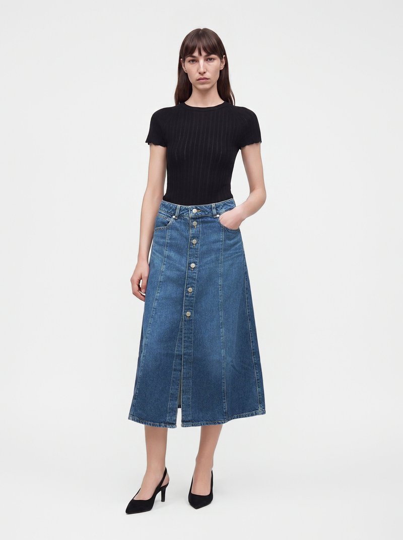 Femme portant un haut noir à manches courtes, une jupe midi en denim bleu boutonnée et des talons noirs, debout devant un fond blanc uni.