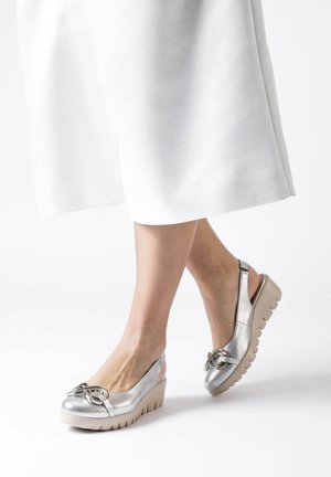 Pies que llevan zapatos con cuña plateados con tira en el talón y cadena metálica decorativa, combinados con una falda midi blanca sobre un fondo liso.