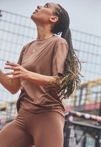 Camiseta atlética marrón con un pequeño logo, combinada con leggings marrones a juego. Presenta un cabello trenzado texturizado; el movimiento sugiere actividad.