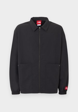 Zwarte jas met ritssluiting en kraag, twee zakken aan de voorkant, elastische manchetten en een klein rood logolabel op de linker mouw.