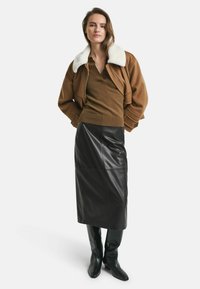 Bruine bomberjack met witte schapenvacht kraag, bruine top, zwarte leren midi rok en zwart kniehoge laarzen, waardoor een gelaagde look ontstaat.