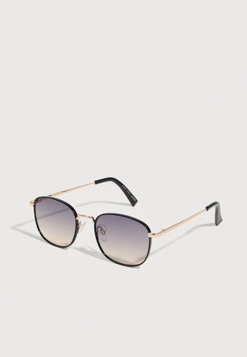 Gafas de sol de forma cuadrada con montura metálica negra y dorada y lentes oscuras con degradado, apoyadas sobre una superficie blanca.