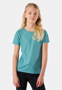 Ung person med langt blondt hår iført en teal kortærmet skjorte og sorte bukser, stående mod en ensfarvet lys baggrund.