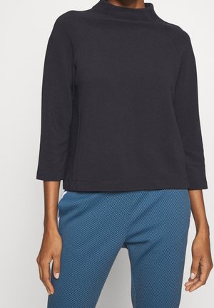 Femme portant un haut noir à manches longues côtelé et un pantalon bleu texturé, debout devant un fond clair uni.