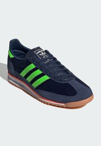 Pantofii sport din suede naval cu trei dungi verzi, talpă din cauciuc și o etichetă logo neagră pe limbă. Dispun de șireturi rotunde și un toc texturat.