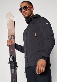 Veste de ski noire avec des accents orange, poche zippée et capuche, tenant un ski marron avec fixations. Des lunettes de soleil réfléchissantes argentées complètent le look.
