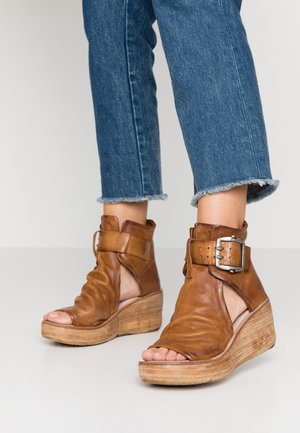 Fødder i cropped blå jeans iført brune læderåbne kilehælssandaler med store spænderem.