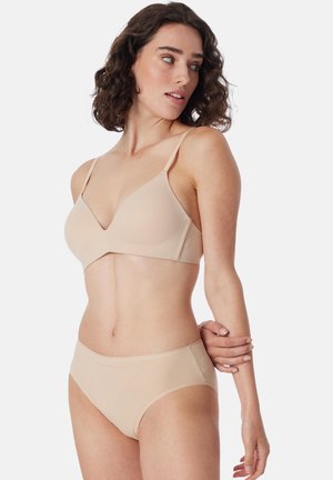 Woman with curly dark hair wearing beige bra and matching underwear, looking to her left, holding her right arm with her left hand.