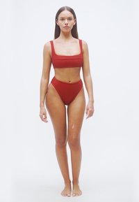 Κόκκινο μπικίνι δύο τεμαχίων με ραβδώσεις, διαθέτει τετράγωνο ντεκολτέ και ψηλόμεσο κάτω μέρος. Το υλικό είναι ελαστικό με λεία υφή.