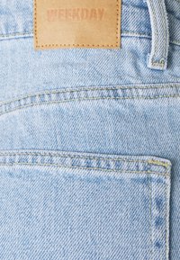 Jasnoniebieskie dżinsy z tylną kieszenią, żółtym szwem i brązową skórzaną metką z wytłoczonym napisem "WEEKDAY".