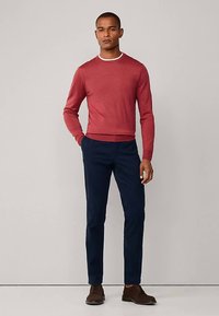 Roter Pullover mit Rundhalsausschnitt und weißem Besatz, kombiniert mit marineblauen Hosen und braunen Schuhen. Der Pullover ist figurbetont, der Stoff wirkt glatt.