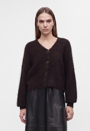 Donna con capelli castani lisci indossa un cardigan marrone scuro soffice con bottoni e una gonna in pelle nera su uno sfondo chiaro uniforme.