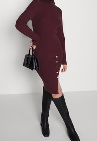 Žena oblečená v těsně přiléhajících bordó šatech z příze s bočními knoflíky a rozparkem, držící černou kabelku a obut�á v černých kozačkách po kolena.
