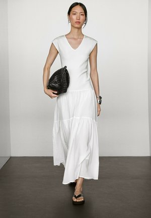 White, cap-sleeve maxi dress with a V-neck and tiered hem; model holds a black, woven handbag and wears black flip-flops.