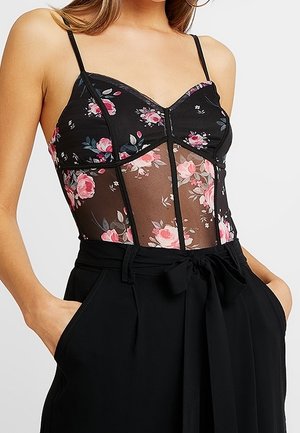 Femme portant un bustier en résille noire à motif floral avec fines bretelles et un pantalon noir taille haute avec ceinture nouée, mains dans les poches.