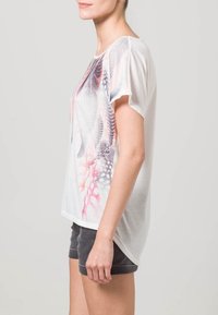 Bílý krátký rukáv tričko s barevným abstraktním designem v růžové a šedé, volný střih, s měkkou texturou. V kombinaci se šedými šortkami.