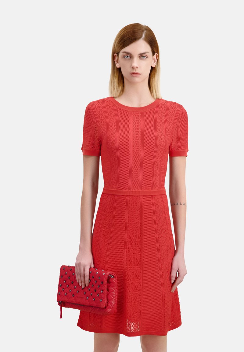 Robe tricotée rouge à manches courtes, avec un motif texturé et une taille cintrée. Associée à une pochette rouge matelassée avec des clous décoratifs.
