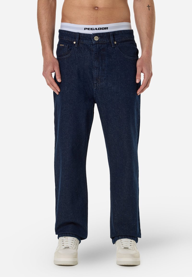 Σκούρα μπλε τζιν π�αντελόνια με ίσια γραμμή, που διαθέτουν πέντε τσέπες, κούμπωμα με κουμπί μπροστά και διακριτή υφή. Συνδυασμένα με άσπρα αθλητικά παπούτσια.