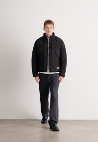 Young man walking indoors wearing a black quilted jacket, light shirt, dark jeans, and black shoes against a white wall background.