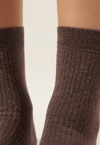 Primo piano di gambe che indossano calzini a coste marrone scuro con fili di glitter argento su uno sfondo chiaro uniforme.