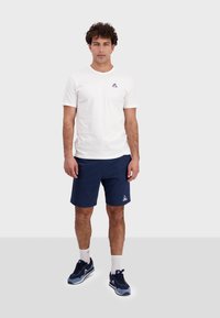 Vit kortärmad t-shirt med en liten logotyp, ihopparad med marinblå shorts. Modellen bär marinblå träningsskor och vita ankelsockor.