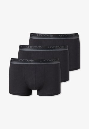 Männlicher Oberkörper, der schwarze Boxer-Briefs mit grauem Bündchen trägt, auf dem das Logo "unc" sichtbar ist und sich um die Taille wiederholt.