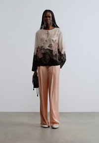 Blouse en soie avec imprimé floral beige et noir, associée à un pantalon en satin rose. Mannequin tenant un petit sac à main noir, portant des chaussures blanches.