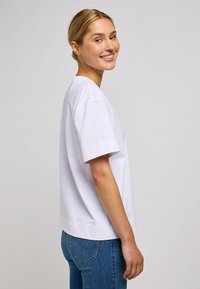 Camiseta de algodón blanca de manga corta, con cuello redondo y un corte relajado. Llevada con jeans de mezclilla azul, vista de perfil.