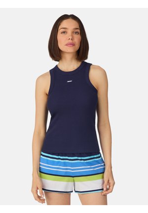 Vrouw met steil, schouderlang bruin haar die een marineblauw mouwloos topje en meerkleurige gestreepte korte broek draagt tegen een effen achtergrond.