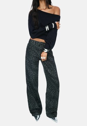 Kobieta w czarnym swetrze odsłaniającym ramiona, ciemnych szerokich spodniach w panterkę, srebrnych szpiczastych butach oraz srebrnych bransoletkach i pierścionkach.