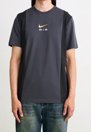 Henkilö, jolla on tumma harmaa Nike Air T-paita kultaisella logolla, yhdistettynä haalistuneisiin sinisiin farkkuihin, seisoo yksivärisen taustan edessä.