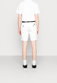 Pantaloni corti bianchi in cotone con una vita nera, dotati di due tasche posteriori con bottoni neri, indossati con una maglietta bianca e scarpe nere.