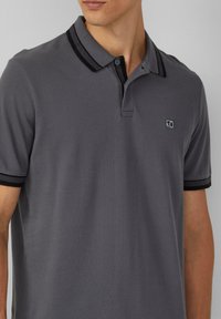 Grijze polo met een textuurafwerking, featuring zwarte accenten op de kraag en de mouwen, een knoopsluiting met twee knopen, en een klein logo op de borst.