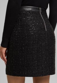 Gros plan sur une mini-jupe noire texturée à sequins, avec une fermeture éclair argentée visible et une ceinture en cuir, portée avec un haut noir à manches longues.
