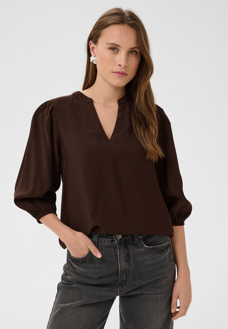 Jeune femme aux longs cheveux bruns portant des boucles d'oreilles argentées, un chemisier ample brun foncé avec des manches bouffantes 3/4, et un jean noir taille haute, une main dans la poche.