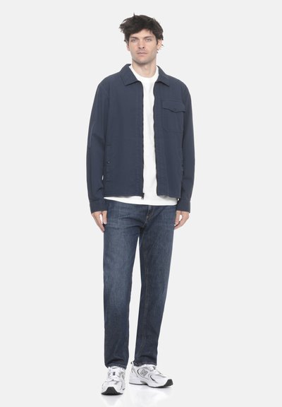 Giovane uomo con giacca blu navy, t-shirt bianca, jeans blu e sneakers bianche in piedi contro uno sfondo bianco uniforme.