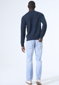 Marineblauwe gebreide trui met lange mouwen, gecombineerd met lichtblauwe jeans. De outfit wordt compleet gemaakt met lichtgekleurde schoenen.