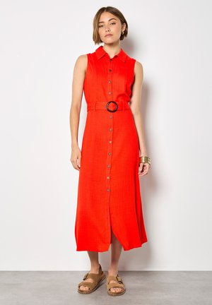 Kobieta w czerwonej, bezrękawnikowej sukience midi z guziczkami z przodu i paskiem, w parze z brązowymi sandałami i złotymi bransoletkami, stoi na tle białej ściany.