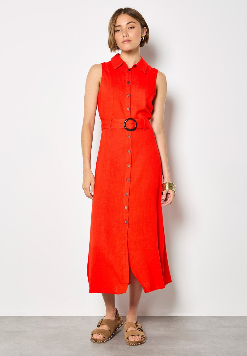 Femme portant une robe midi rouge sans manches avec des boutons sur le devant et une ceinture, associée à des sandales beiges et des bracelets dorés, debout contre un mur blanc.